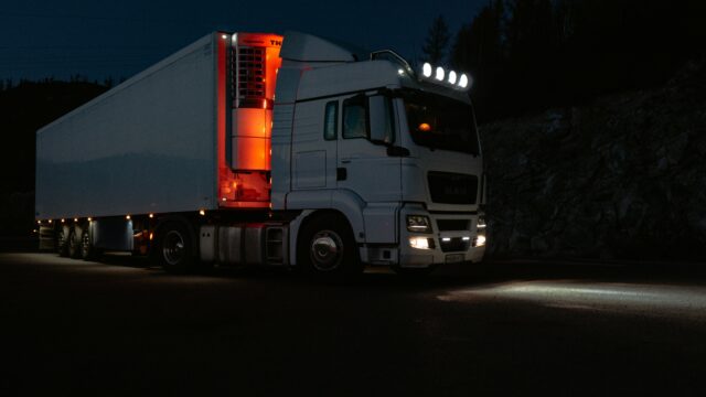 A white truck lit by headlights and interior lights on a dark road in mountainous terrain.