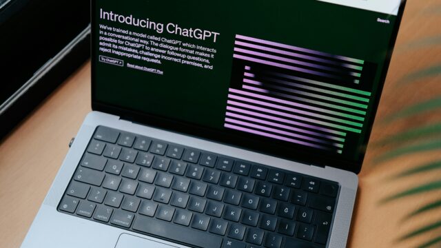 A sleek laptop on a wooden desk showing the ChatGPT homepage by a window.