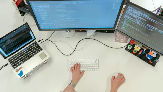 A female software engineer coding on dual monitors and a laptop in an office setting.