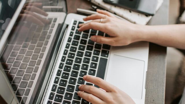 From above of unrecognizable woman sitting at table and typing on keyboard of computer during remote work in modern workspace