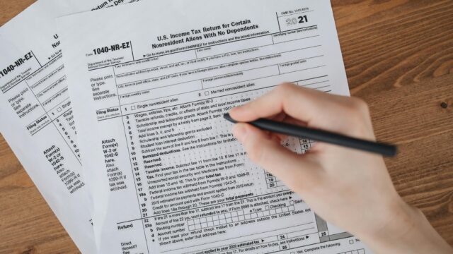 Person filling out U.S. tax form 1040-NR-EZ with a pen on a wooden table.