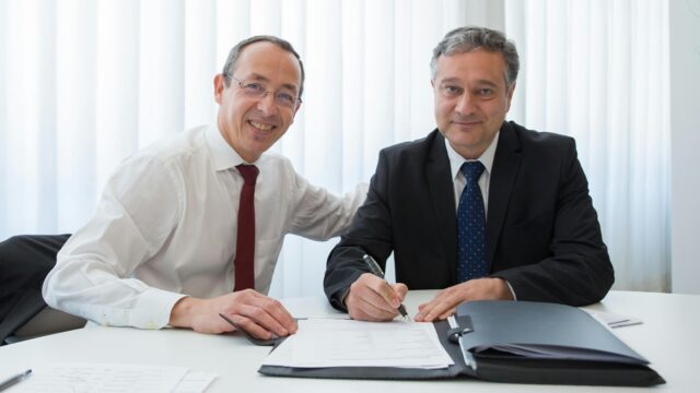 Two businessmen in suits signing a contract at a well-lit office table.