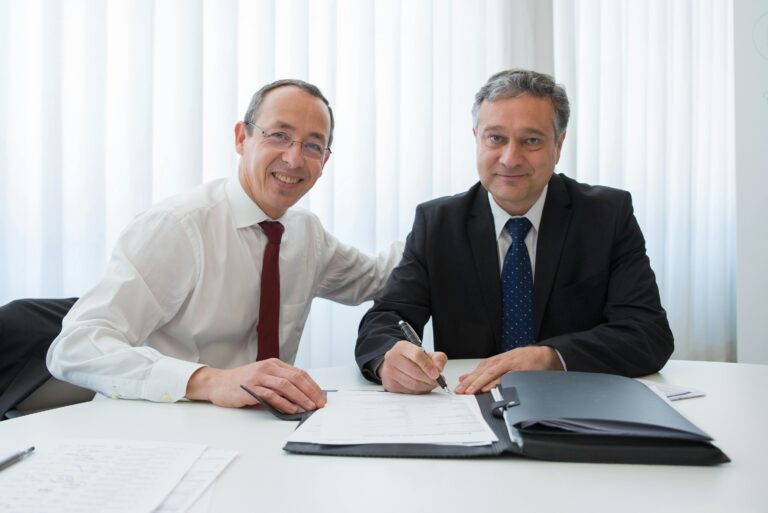 Two businessmen in suits signing a contract at a well-lit office table.