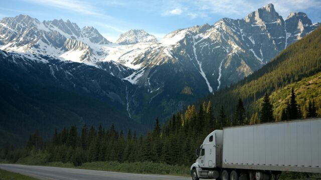 A semi-truck travels along a highway with snow-capped mountains in the background.